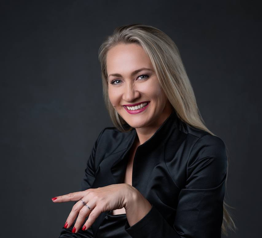 Kate Kharlamova seated in a black satin dress, smiling against a dark studio backdrop