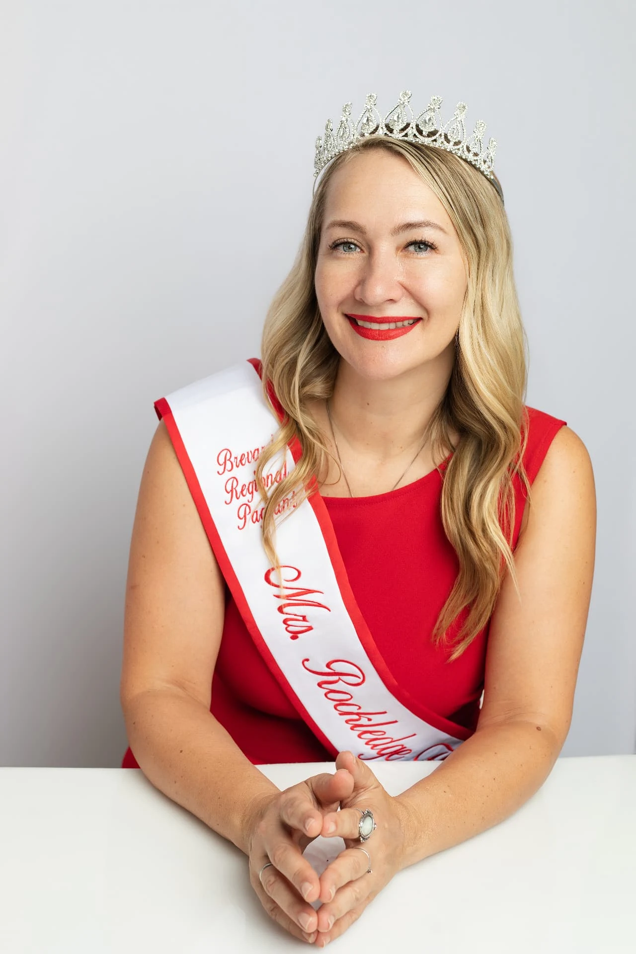 Kate Kharlamova in her red dress and Mrs. Rockledge sash, hands clasped, against a light studio backdrop